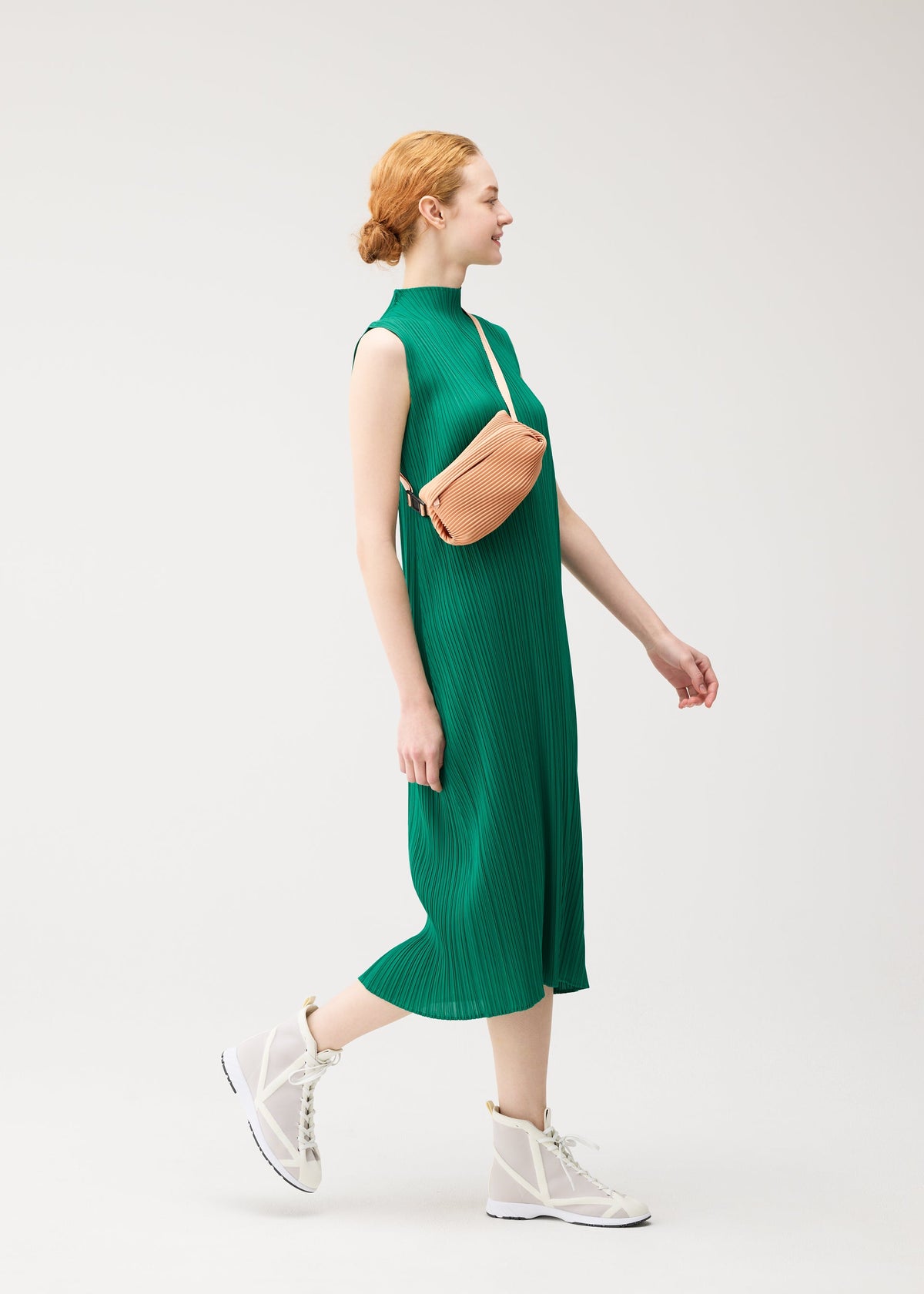 A woman in a sleeveless green BIAS PLEATS dress by PLEATS PLEASE ISSEY MIYAKE, white mesh sneakers, and a light tan WAIST BAG walks against a plain white background, smiling and looking to the side. view 6 of 7
