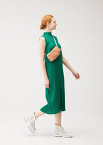 A woman in a sleeveless green BIAS PLEATS dress by PLEATS PLEASE ISSEY MIYAKE, white mesh sneakers, and a light tan WAIST BAG walks against a plain white background, smiling and looking to the side.