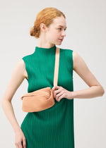 A woman in a sleeveless green pleated dress holds the BIAS PLEATS WAIST BAG by PLEATS PLEASE ISSEY MIYAKE, a peach-hued crossbody with diagonal pleats, adding a chic accent against a simple light background.