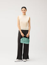 A woman stands against a plain light background, wearing a sleeveless cream top and black pleated pants with bold diagonal pleating. She holds the BIAS PLEATS BAG by PLEATS PLEASE ISSEY MIYAKE—a small green shoulder bag—and smiles softly at the camera, completing her look with white sneakers.