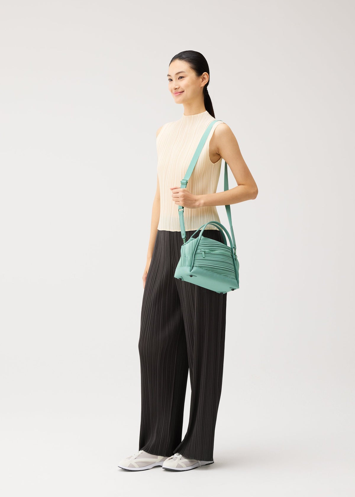 A woman stands smiling, wearing a sleeveless beige top, wide-leg black pants, white sneakers, and holding the BIAS PLEATS BAG by PLEATS PLEASE ISSEY MIYAKE—a small teal bag featuring elegant diagonal pleating. The background is plain and light. view 10 of 10