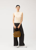A woman stands smiling, dressed in a sleeveless cream top, wide-leg black pants, and white sneakers. She holds the BIAS PLEATS BAG by PLEATS PLEASE ISSEY MIYAKE with both hands; its brown textured surface adds subtle elegance to her look. The background is plain and light-colored.