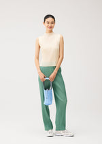 A woman stands smiling against a plain light background, dressed in a sleeveless cream top, green pleated pants, white sneakers, and carrying the PLEATS SWAN BAG by PLEATS PLEASE ISSEY MIYAKE featuring an elegant swan motif.