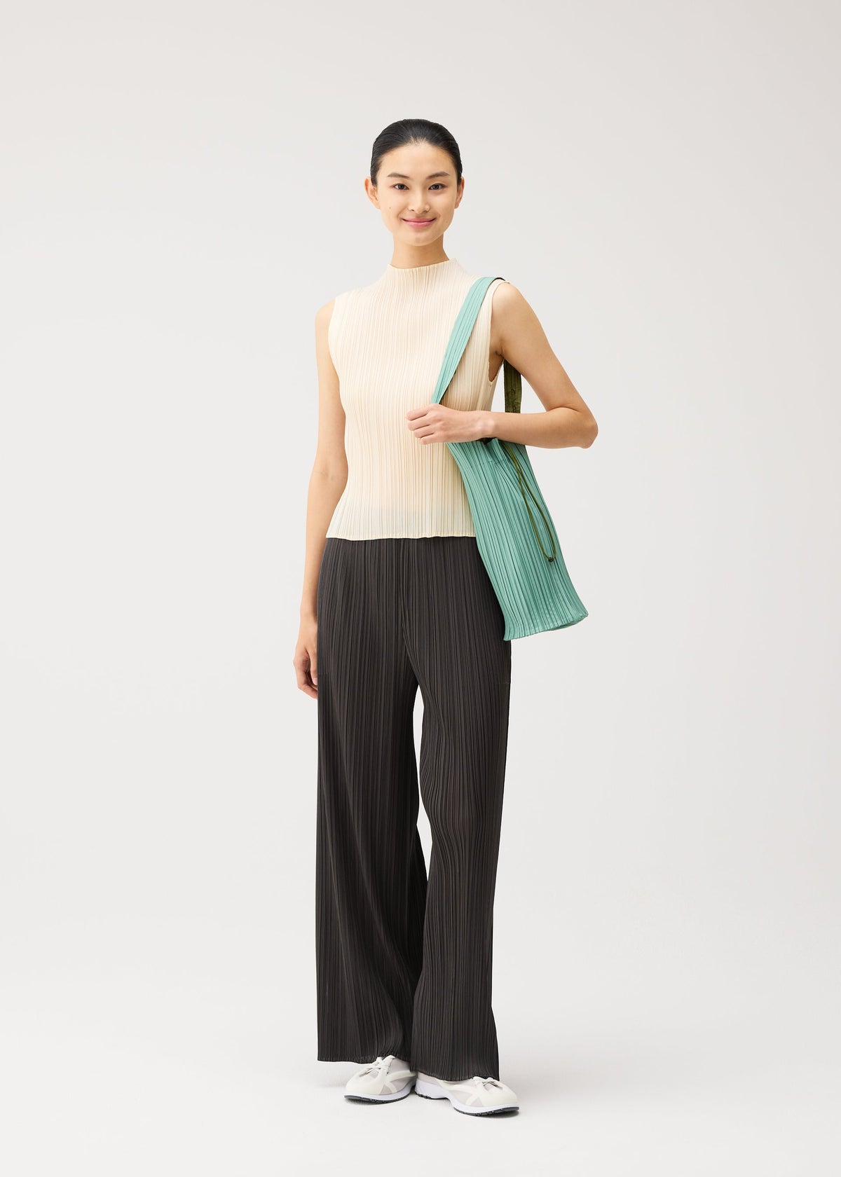 A woman stands against a plain background, wearing a sleeveless beige top, loose black pinstripe pants, and white sneakers. She has her hair tied back in a slight smile, and carries the elegant PLEATS SWAN BAG by PLEATS PLEASE ISSEY MIYAKE over her shoulder. view 6 of 6