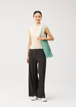 A woman stands against a plain background, wearing a sleeveless beige top, loose black pinstripe pants, and white sneakers. She has her hair tied back in a slight smile, and carries the elegant PLEATS SWAN BAG by PLEATS PLEASE ISSEY MIYAKE over her shoulder.
