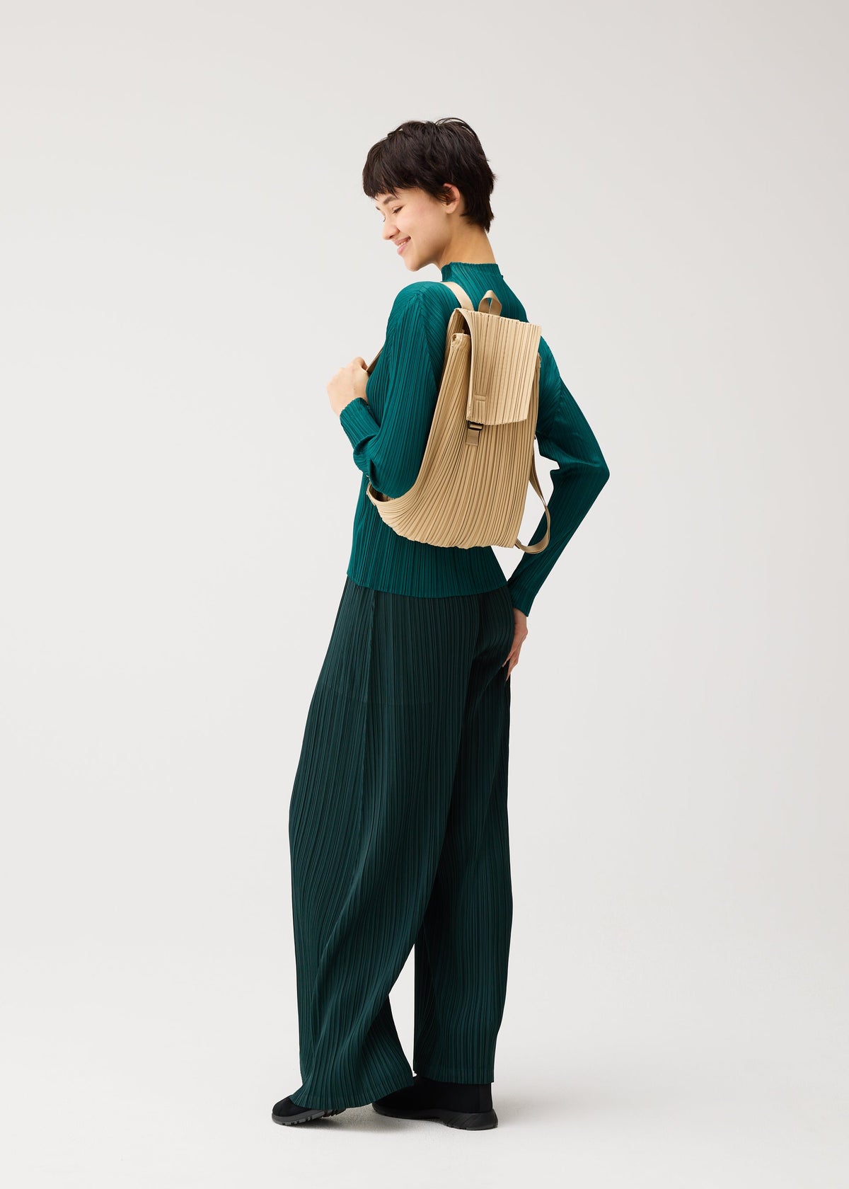 Zoomed view Person in a green pleated outfit and black shoes smiles over their shoulder, carrying the PLEATS KNAPSACK by PLEATS PLEASE ISSEY MIYAKE with a beige textured finish against a plain white background.