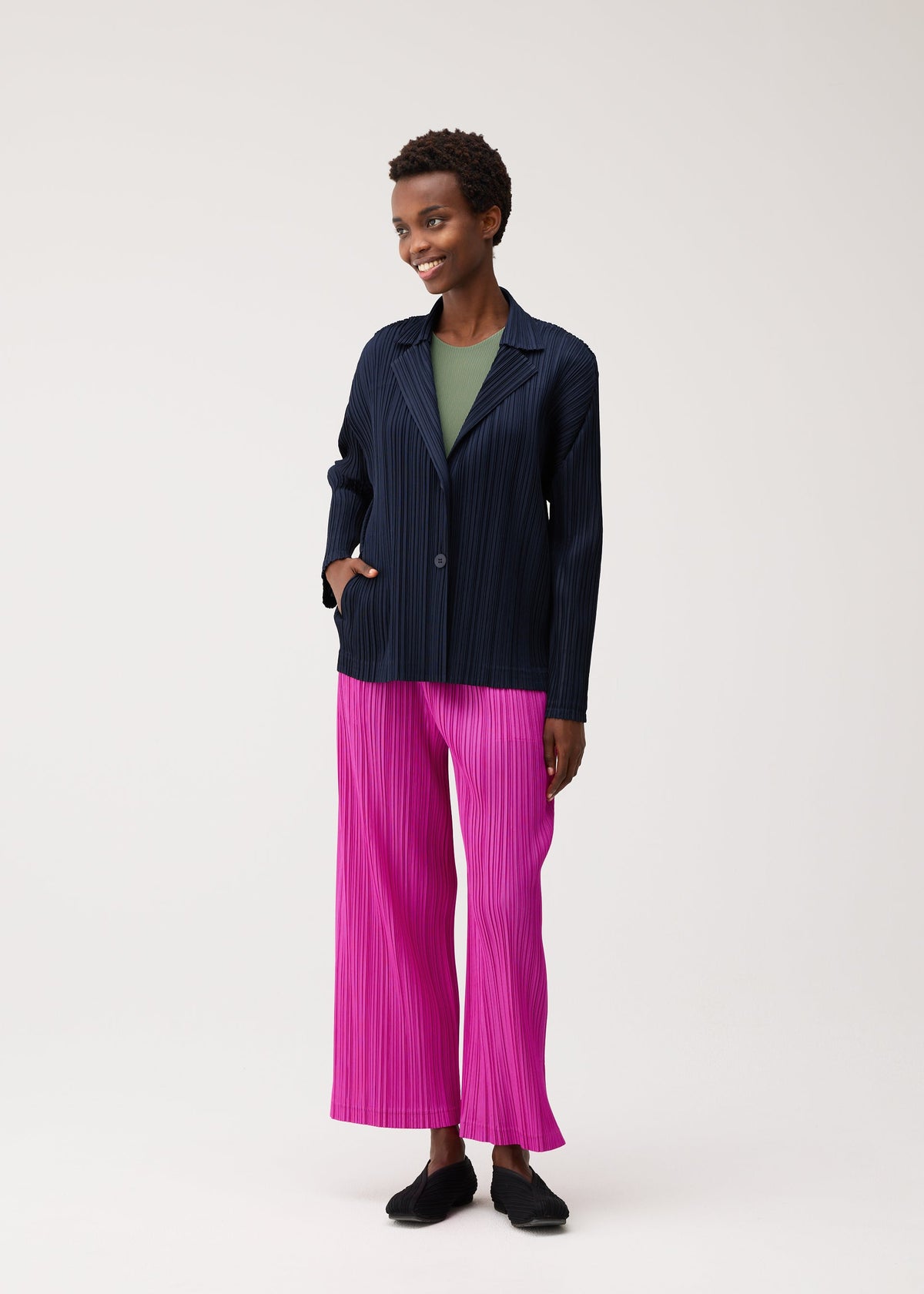 A person stands smiling against a plain light background, wearing a navy pinstripe blazer over a green top, bright pink pleated wide-leg pants, and black KNIT BALLET SHOES by PLEATS PLEASE ISSEY MIYAKE. view 4 of 4
