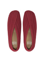 A pair of red KNIT BALLET SHOES by PLEATS PLEASE ISSEY MIYAKE, seen from above, showcase a pleated, knitted fabric texture and a subtly pointed toe. The insoles are marked with “PLATS PLEASE” in white lettering.