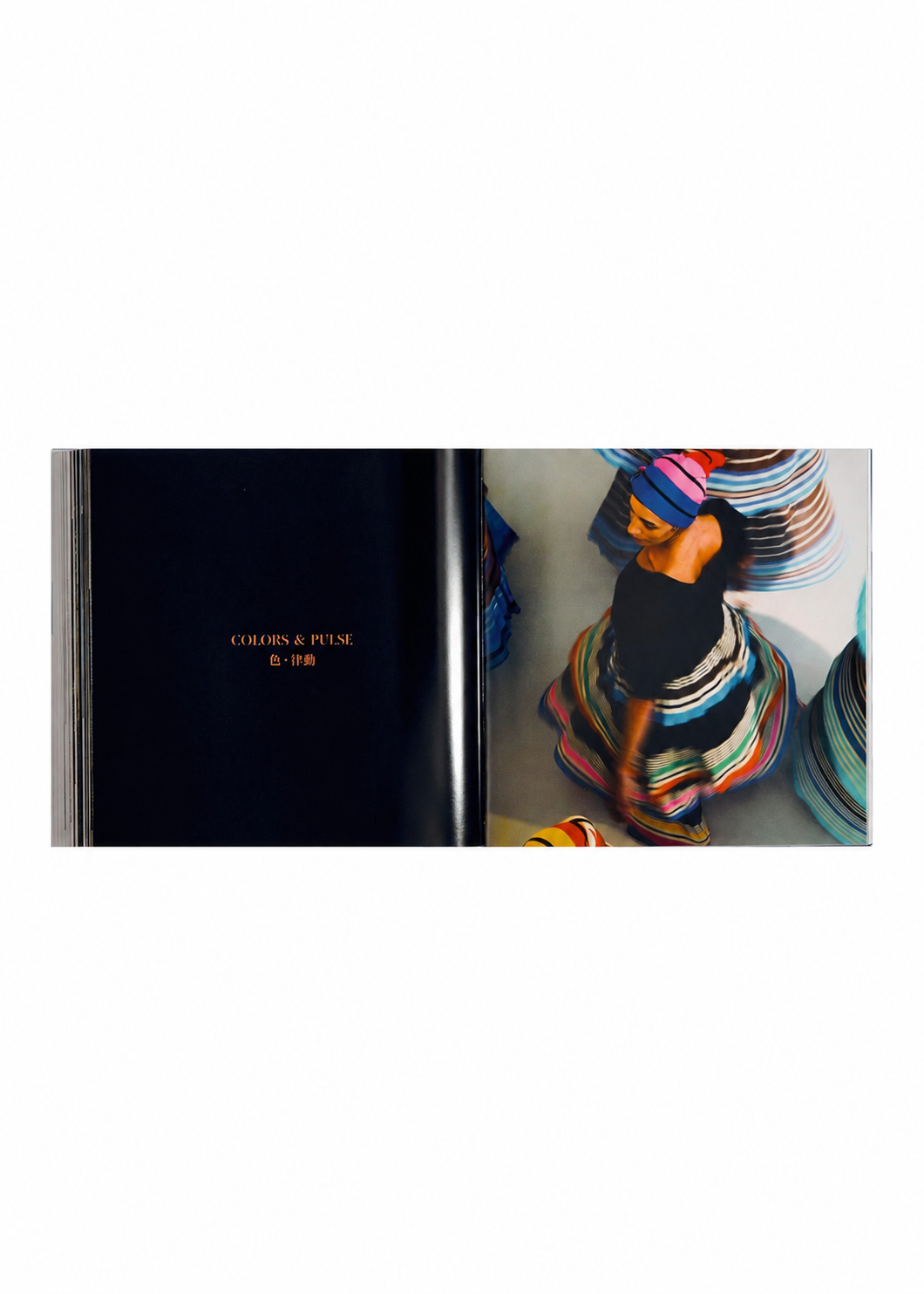 Zoomed view Open book with the left page displaying the words “COLORS & PULSE” in gold text, and the right page featuring a woman in a vibrant, swirling skirt and headwrap, seen from above against a light backdrop—an image that captures the spirit of ISSEY MIYAKE’s design philosophy and artistic innovation. From ISSEY MIYAKE (Limited Collectors Edition) by ISSEY MIYAKE BOOKS.