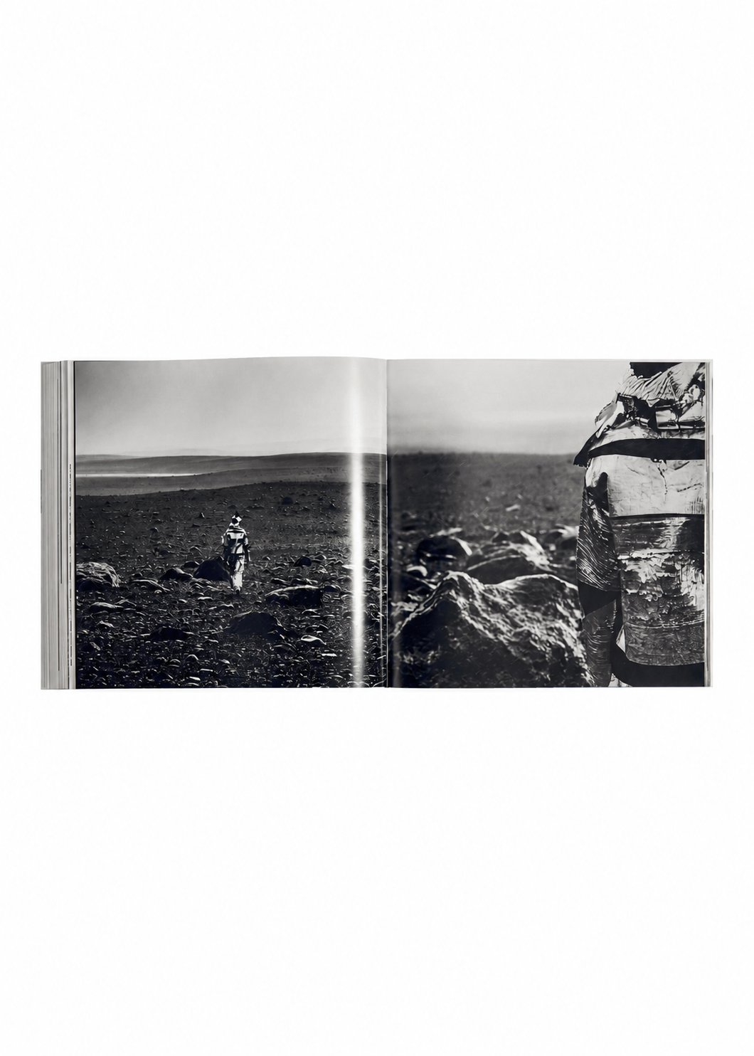 Open book from ISSEY MIYAKE (Limited Collectors Edition) by ISSEY MIYAKE BOOKS, featuring a black-and-white photograph of a stark, rocky landscape. On the left page, a solitary figure stands in the distance, while another person—partially seen and turned away—appears on the right page. The composition echoes Issey Miyake’s distinctive design philosophy and invites thoughtful contemplation, making this edition a striking Collector’s piece. view 13 of 17