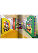 Vivid, cartoon-inspired paintings with bold, rounded frames grace the walls of a luminous, contemporary art gallery featuring yellow and multicolored floors, capturing the lively essence of Tomokazu Matsuyama’s art showcased in “Matsuyama Tomokazu: FIRST LAST” by ISSEY MIYAKE BOOKS.